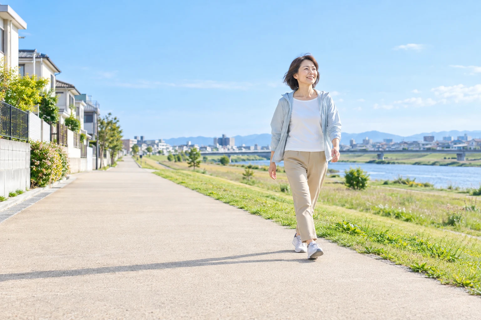 青空の広がる住宅地と川沿いの道を、朝の光の中で歩く女性